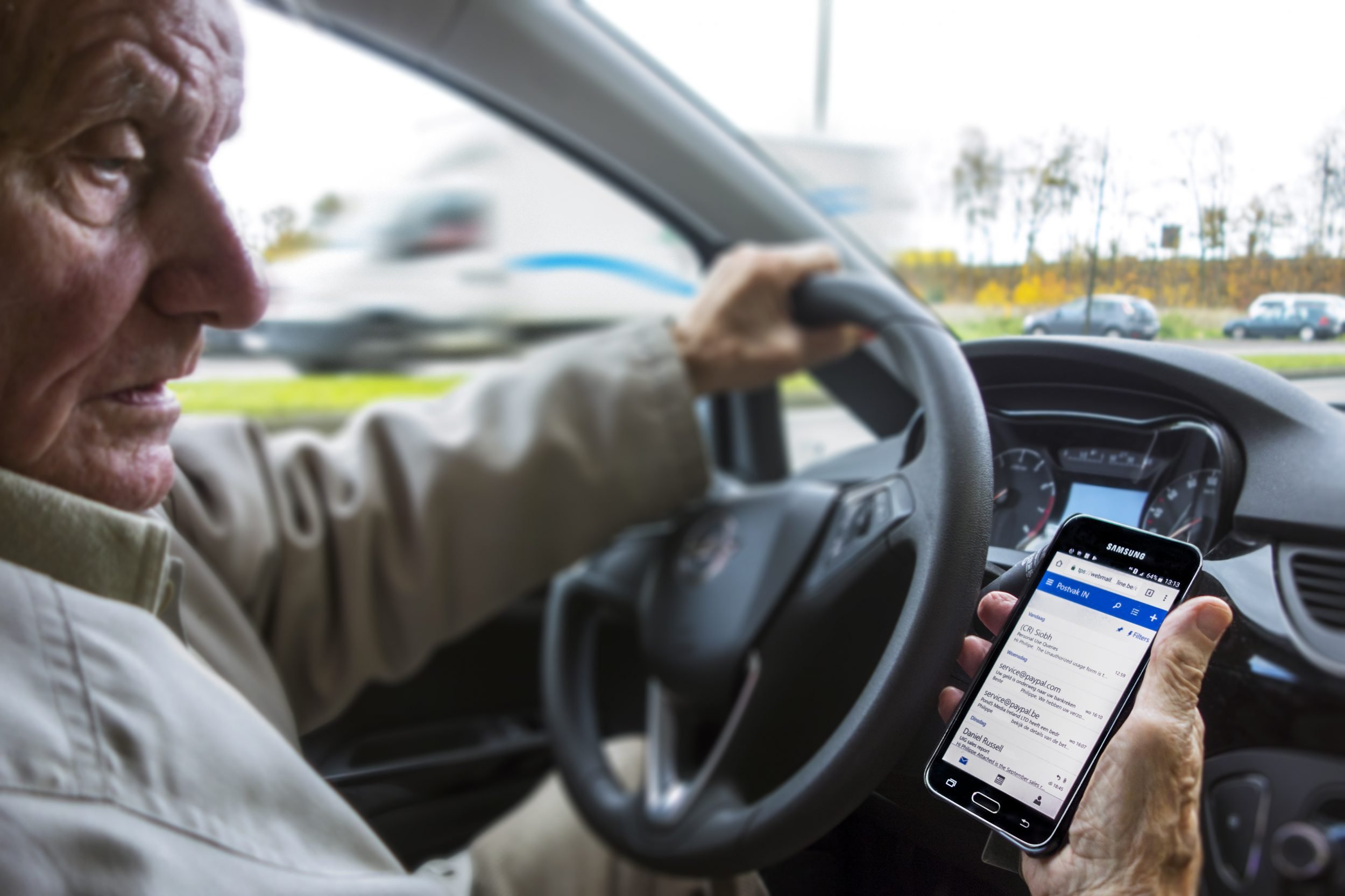 Irresponsible elderly man at steering wheel checking messages on smart phone while driving car on road.